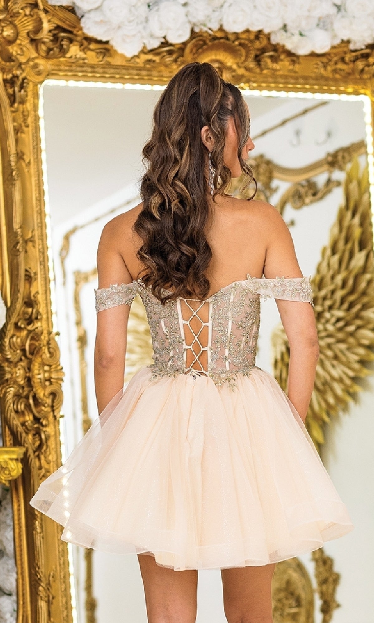 A woman with long, wavy hair stands before a gold ornate mirror in the Off-Shoulder Short Prom Dress: Dancing Queen 3418—an elegant, lace-up back dress that’s stylish, affordable, and perfect for any special occasion.
